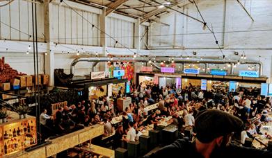 Small coloured food vendors inside an airy warehouse. There is festoon lighting and people sat at tables and benches.