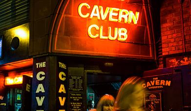 The exterior and entrance of the Cavern Club. The neon red entry sign reads 'Cavern Club.' There are two women leaving the club sminling.