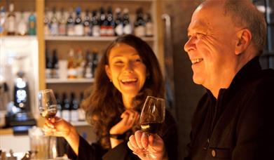A man and woman sitting smiling and holding a small glass of red wine.