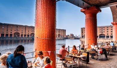 People sitting outside at the Royal Albert Dock. There are large red columns supporting the old warehouse buildings  of the dock.