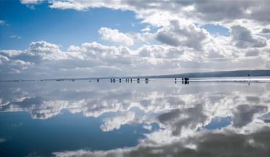 A vast marine lake on the coastline with people walking alongside. The sky is blue with some clouds that are reflecting in the water
